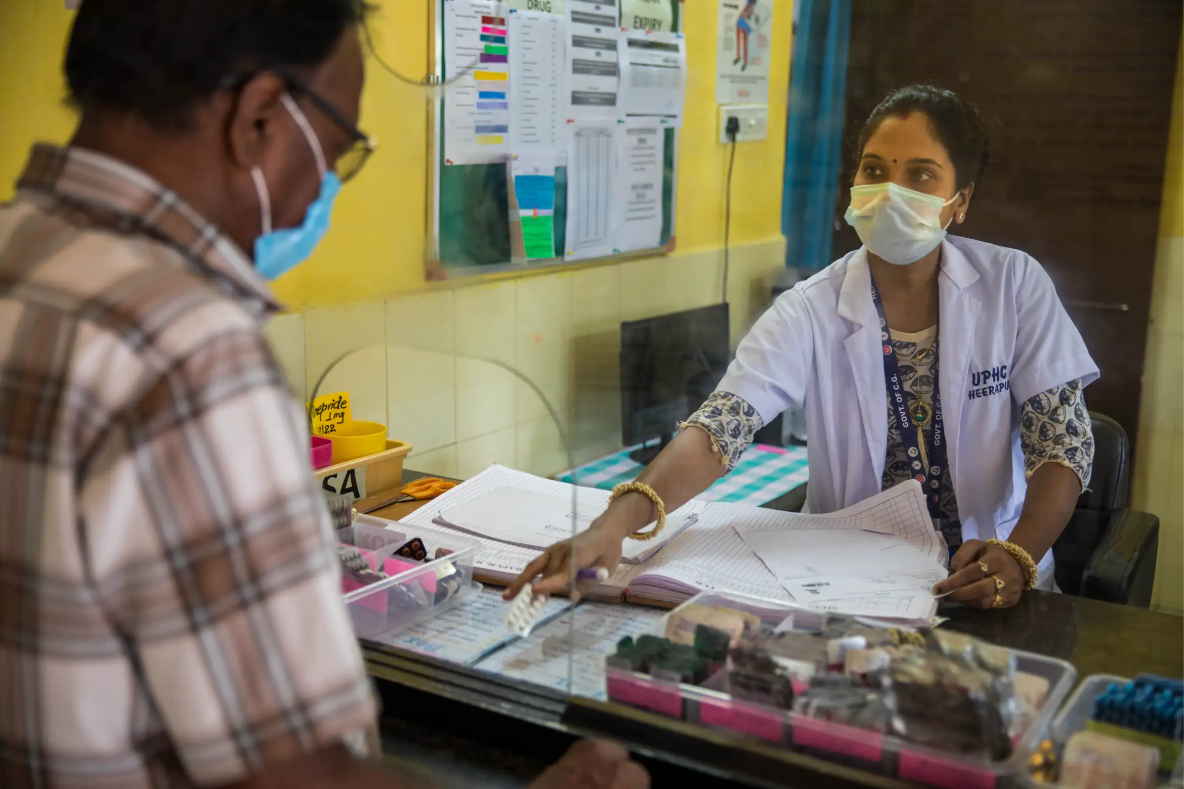 A female Indian health care worker hands a patient a strip of medication through a hole in a protective screen