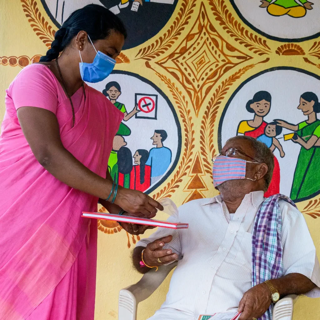 A patient wearing a mask, and sitting in a plastic chair, while a woman in a sari stands next to him, against a brightly coloured mural on the wall.